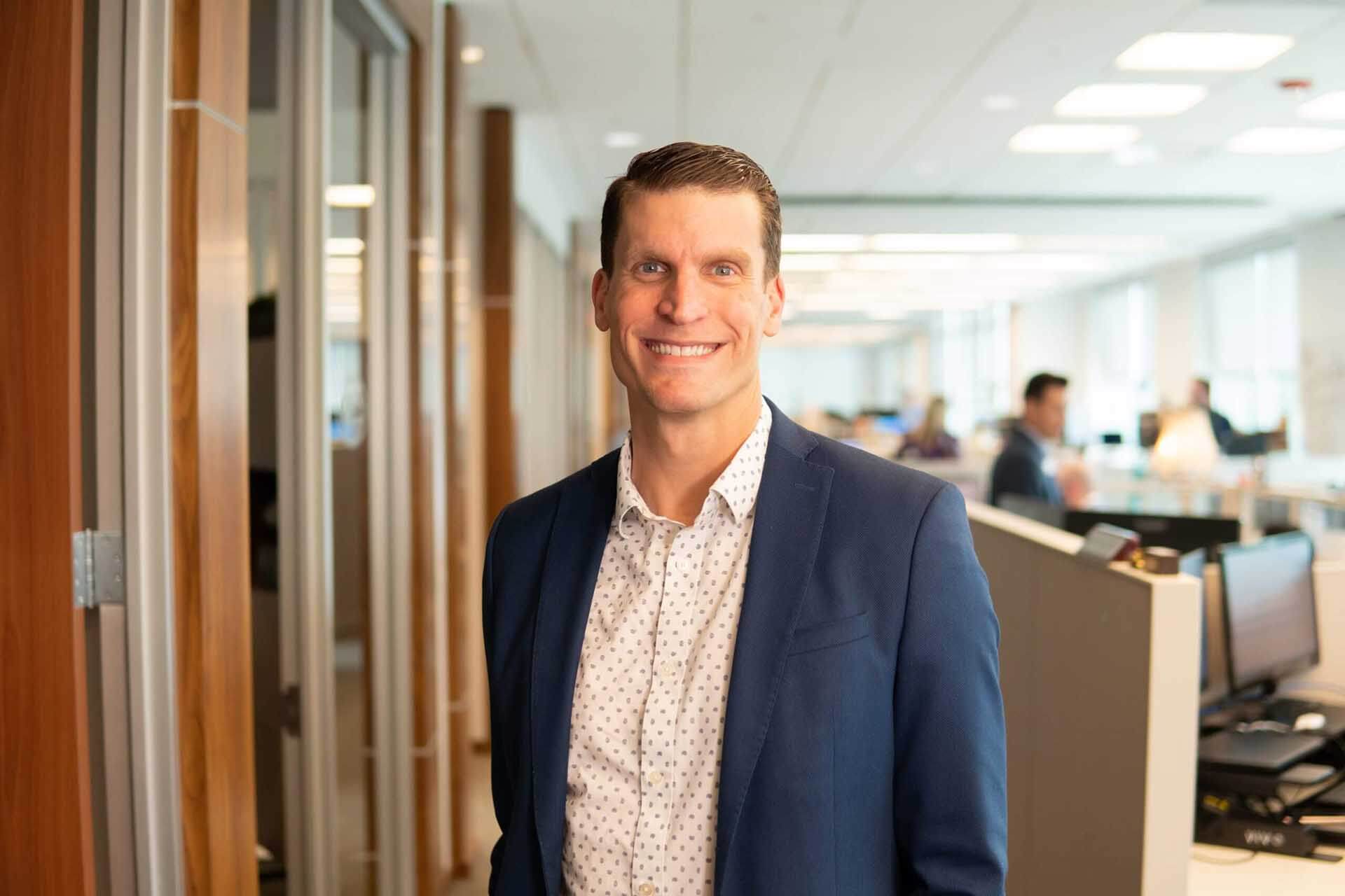 A person in business attire smiles in a modern office environment with people working in the background.